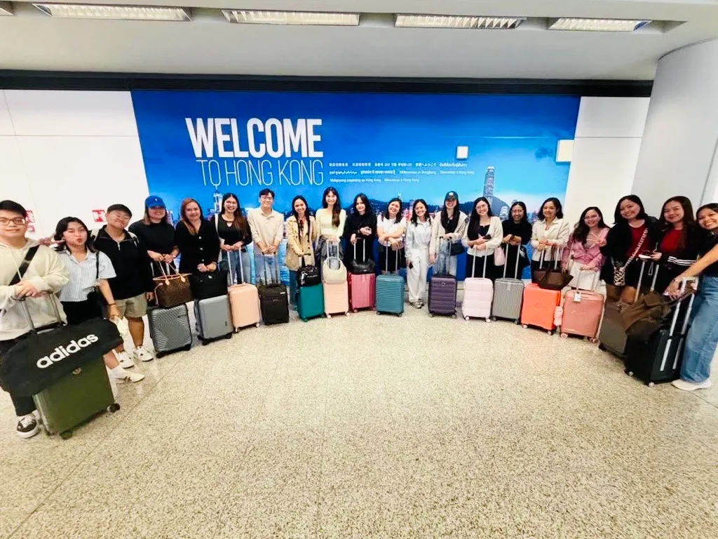 A large group of TeemCorp employees, all of Filipino heritage, happily standing in a row and smiling with their suitcases. They are posed in front of a blue airport welcome sign that reads 'WELCOME TO HONG KONG' with the city skyline, demonstrating a company team-building trip and investment in people.